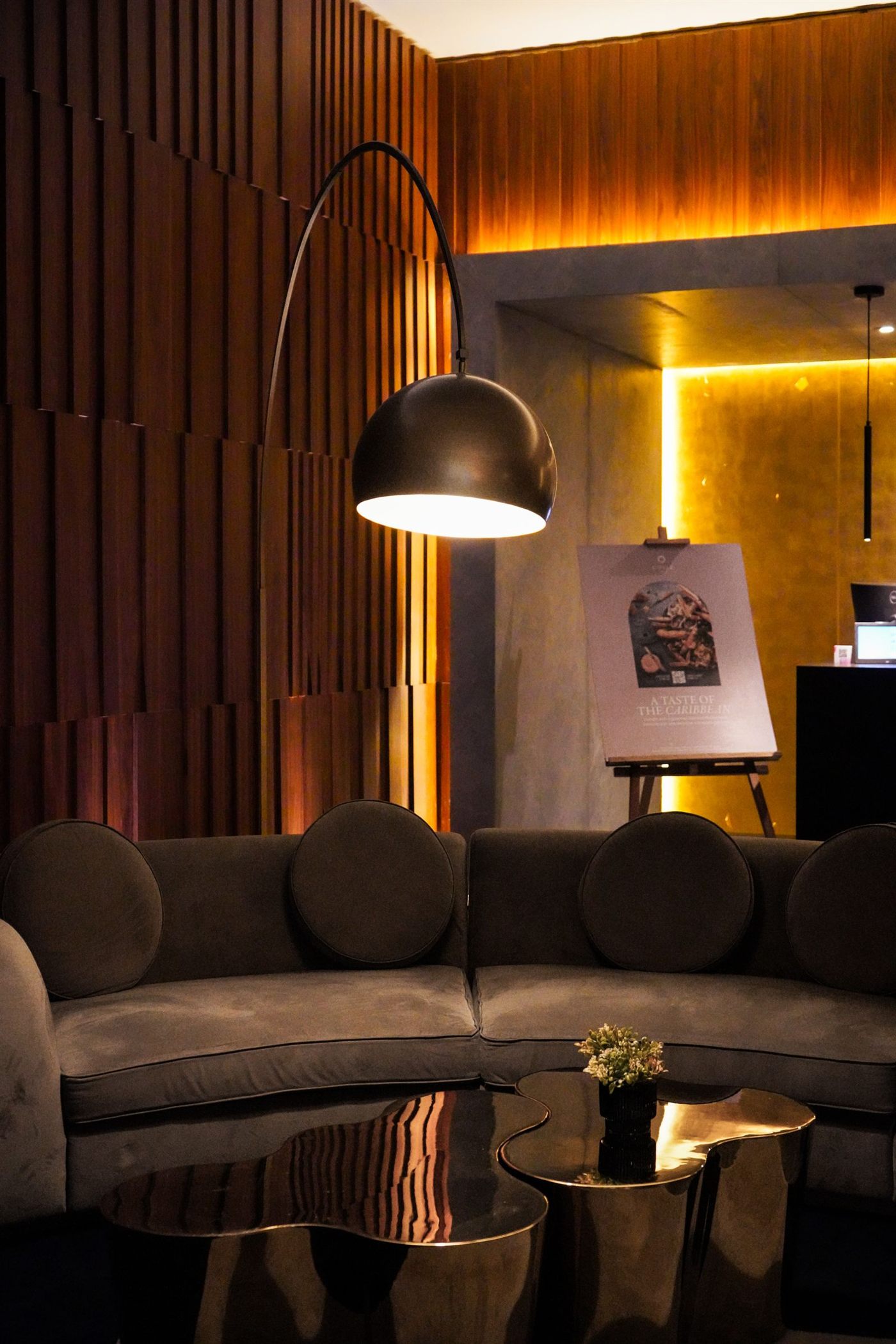 Lounge de hotel en penumbra con muro de listones de madera, sofá de terciopelo café y mesas de latón